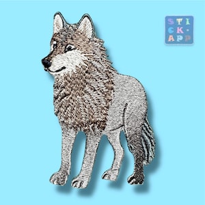 Könnte beinhalten: Gestickter Aufnäher eines stehenden Wolfes mit detailliertem Fell in Braun-, Grau- und Weißtönen. Der Wolf ist vor einem hellblauen Hintergrund platziert. Ein kleines quadratisches Logo mit dem Text "STICK APP" befindet sich oben rechts.