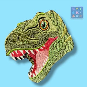 Könnte beinhalten: Gestickter Aufnäher mit einem grünen Tyrannosaurus Rex-Kopf mit offenem Maul, das rote und weiße Zähne zeigt. Der Dinosaurier hat ein detailliertes, strukturiertes Aussehen vor einem hellblauen Hintergrund. Der Aufnäher enthält den Text "STICK APP".