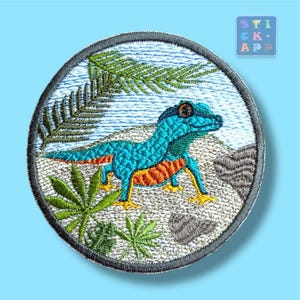 Könnte beinhalten: Runder, gestickter Aufnäher mit einem türkisfarbenen Gecko mit orangefarbenen und gelben Details, vor dem Hintergrund von Farnen und Felsen. Der Aufnäher hat einen grauen Rand und den Text "STICK APP".