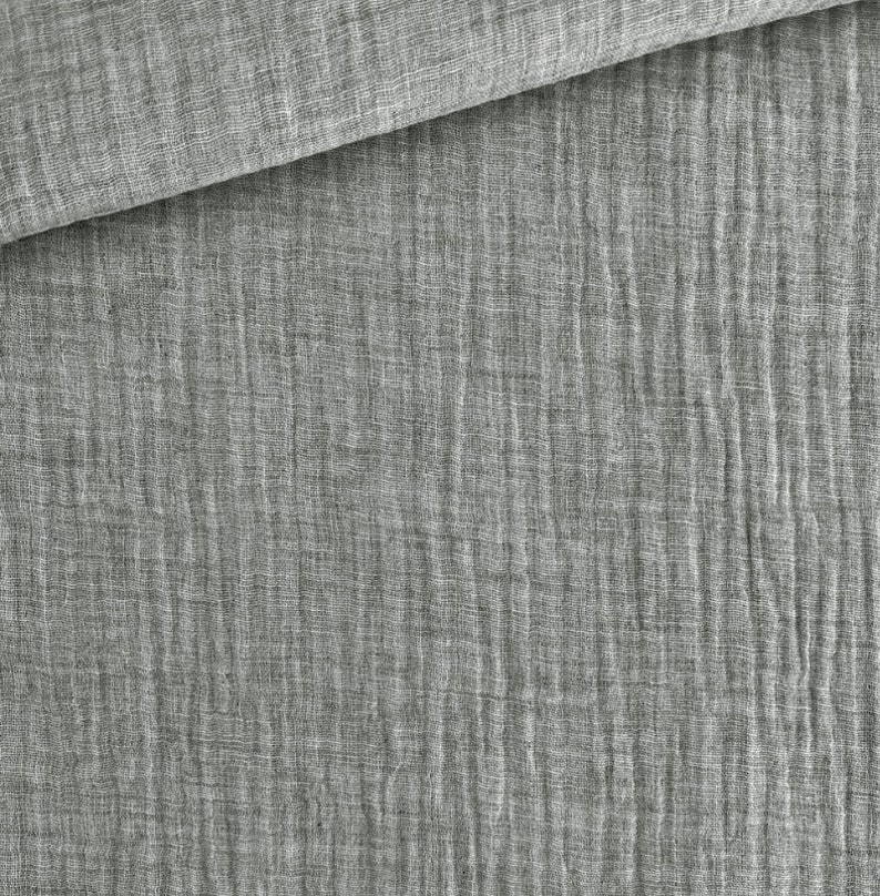 May include: Close-up of a gray fabric with a textured, crinkled appearance. The fabric is folded over, revealing a layered effect. The material has a soft, slightly wrinkled look, suggesting a lightweight textile.