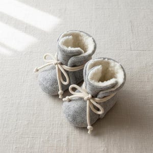 Può includere: Un paio di stivaletti grigi per bambini con un interno bianco morbido e soffice. Gli stivaletti sono legati con uno spago color crema e sono realizzati in un materiale morbido simile al feltro. Gli stivaletti sono progettati per tenere caldi i piedi del bambino.