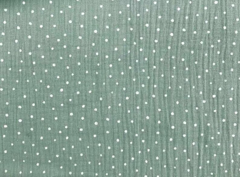 May include: Close-up of a textured fabric in a muted green color, scattered with small, evenly spaced white polka dots. The fabric has a slightly crinkled appearance, suggesting a soft, breathable material. Ideal for apparel or home decor.