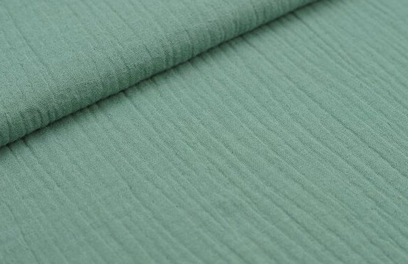 May include: Close-up of a folded piece of sage green fabric. The fabric has a textured, ribbed appearance, with vertical lines running across the material. The fabric appears soft and lightweight, suitable for various sewing projects.