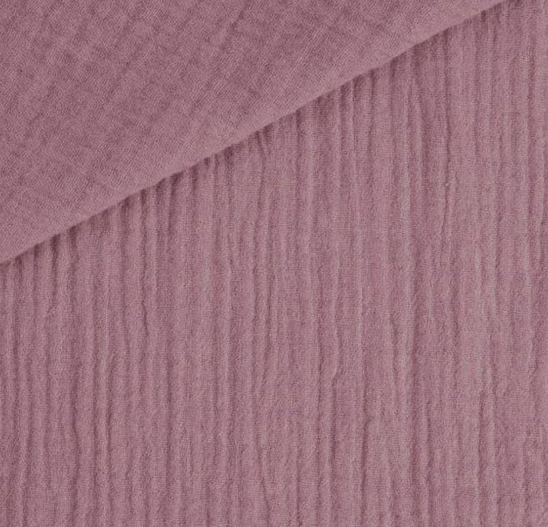 May include: Close-up of a dusty rose-colored fabric with a crinkled texture. The fabric is folded diagonally, revealing a layered effect. The material has a soft, matte appearance, suitable for various textile projects.