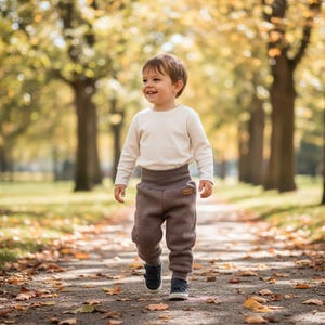 Pode incluir: Uma criança pequena veste uma camisola de manga comprida cor de creme e calças de mistura de lã castanha com uma cintura alta e larga. As calças têm uma pequena etiqueta retangular castanha na anca e ténis azul-escuros com velcro.