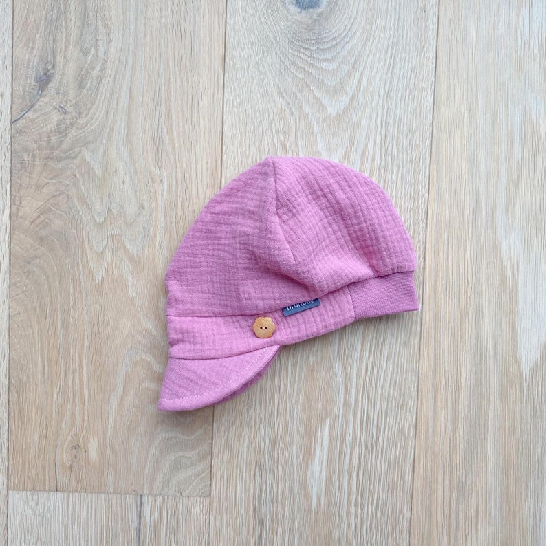 May include: A light pink baby hat with a small brim and a button detail. The hat is made of a textured fabric and features a small label with text. The hat is designed for infants and toddlers.