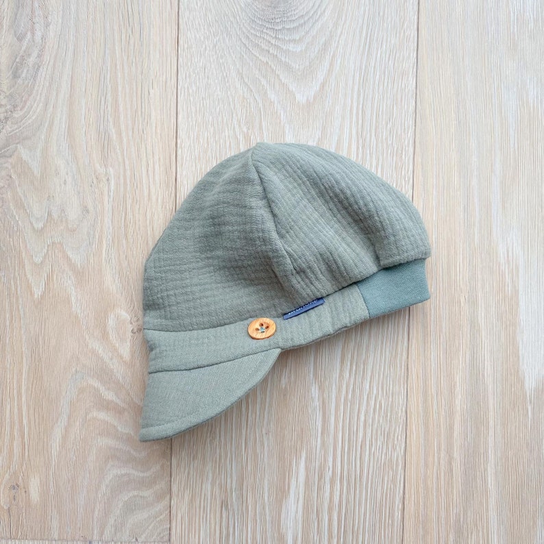 May include: A sage green baby hat with a small brim and a button detail. The hat is made of a textured fabric and has a contrasting green band around the bottom. A small blue tag is sewn on the side.