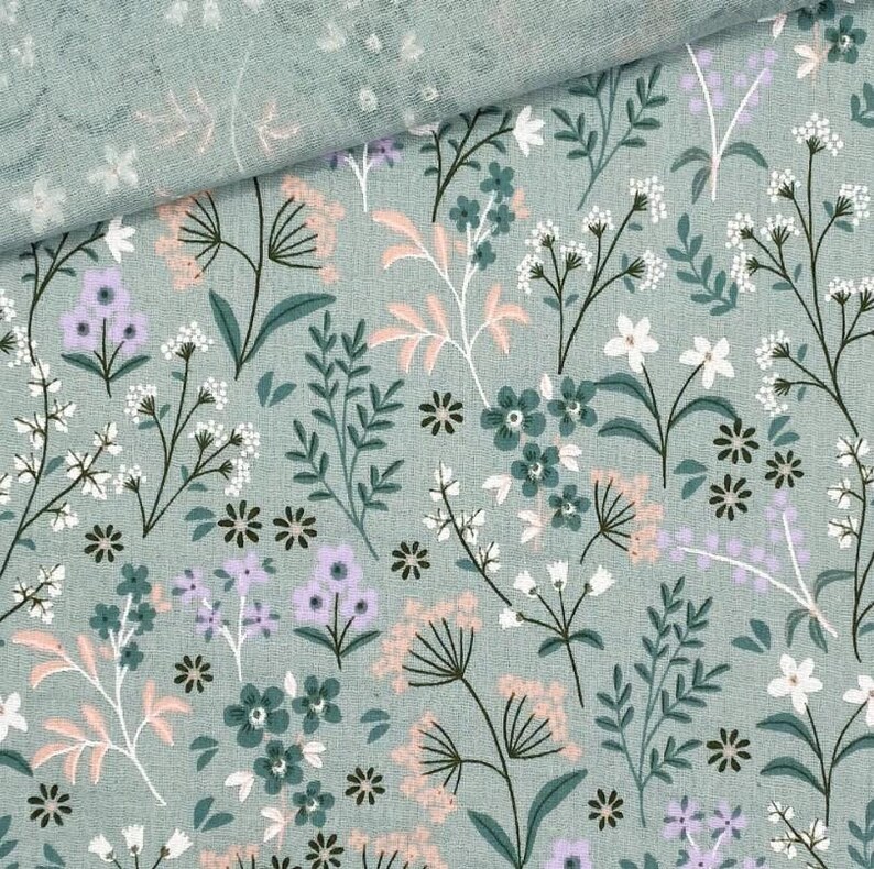 May include: A close-up of a fabric with a floral pattern. The fabric is a light green color with a repeating pattern of flowers and leaves in shades of white, pink, purple, and green. The fabric is folded over in the upper left corner.