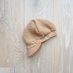 May include: A tan baby hat with a small brim and ear flaps, featuring a wooden button closure. The hat has a soft, textured appearance and a small blue tag with the brand name.
