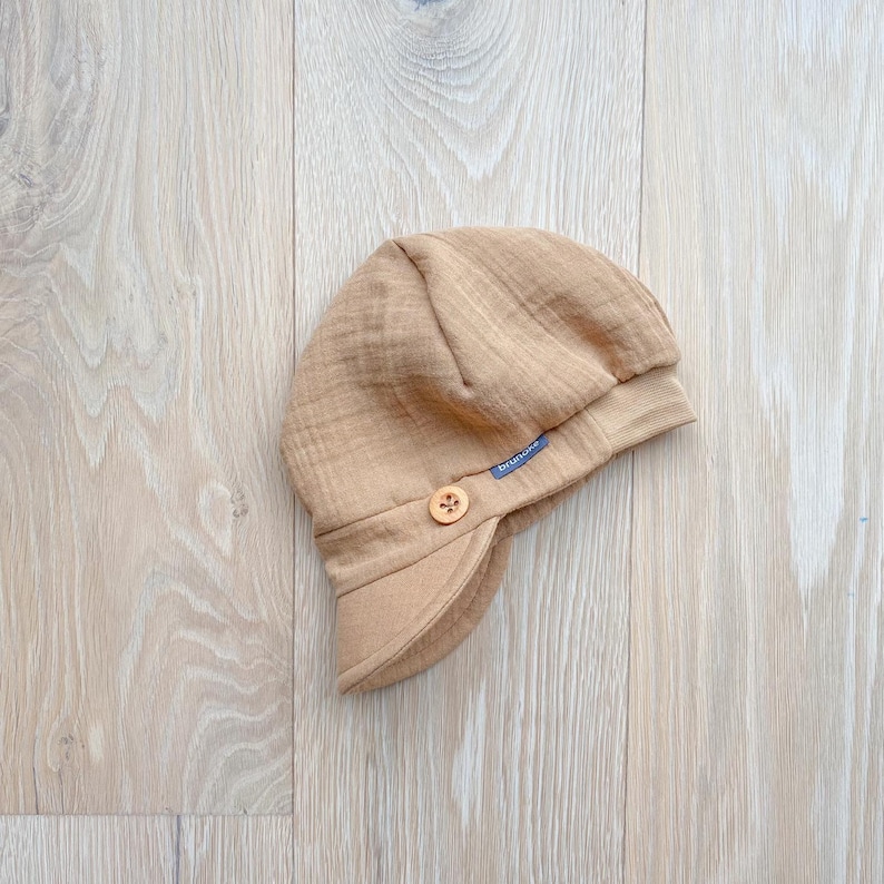 May include: A tan baby hat with a small brim and ear flaps, featuring a wooden button closure. The hat has a soft, textured appearance and a small blue tag with the brand name.