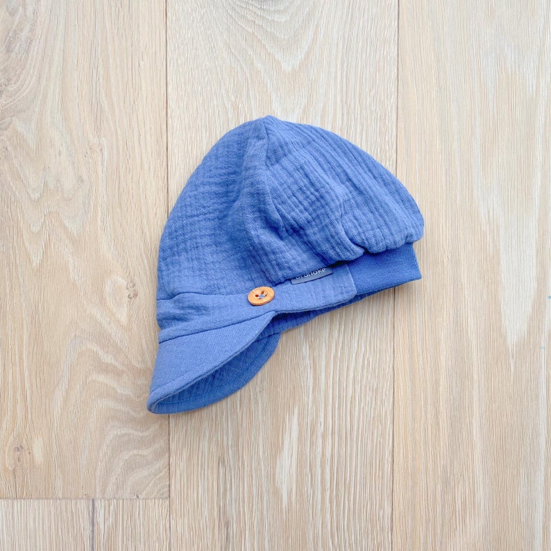 May include: A blue baby hat with a brim and a wooden button closure. The hat is made of a textured fabric and has a rounded crown. The brim is a darker shade of blue. The hat is on a light wood surface.