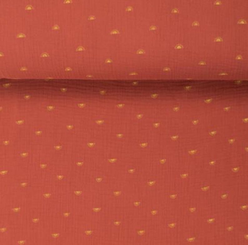 May include: Close-up of a terracotta-colored fabric with a repeating pattern of small, golden yellow half-circle shapes. The fabric is folded, showing a textured surface. The design is evenly spaced across the material, creating a consistent visual texture.