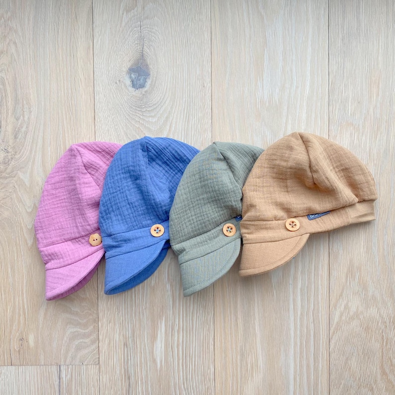May include: Four baby sun hats in pastel colors: pink, blue, green, and tan. Each hat features a small brim and a wooden button detail. The hats are made of a textured fabric and are arranged on a light wood surface.
