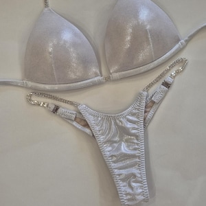 Puede incluir: Conjunto de bikini plateado con top triangular y braguita tanga. El bikini presenta tirantes de pedrería y una tela brillante. El conjunto se muestra sobre una superficie blanca.