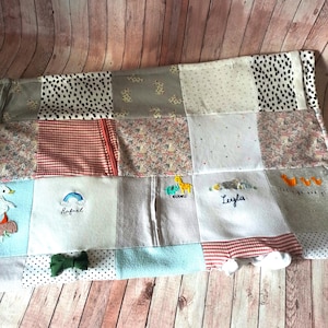 Könnte beinhalten: Eine Patchwork-Babydecke mit verschiedenen Stoffen und Farben. Die Decke hat ein hellblaues Quadrat mit einem weißen Einhorn, ein weißes Quadrat mit einem Regenbogen und dem Namen "Rafael", ein graues Quadrat mit dem Namen "Leyla" und ein graues Quadrat mit dem Namen "Adorable" und einer Giraffe. Die Decke hat auch eine grüne Schleife.