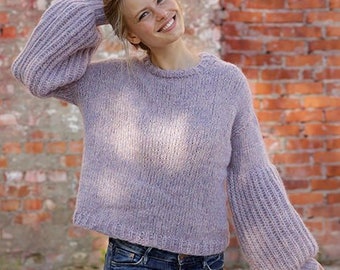 May include: A woman wearing a light purple knitted sweater with a round neckline and long sleeves. The sweater has a textured pattern and a slightly cropped length. The woman is smiling and looking at the camera. The sweater is from DROPS DESIGN. www.garnstudio.com