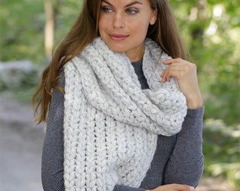 Può includere: Una donna indossa un maglione grigio e una sciarpa di lana bianca. La sciarpa è un punto grosso con un motivo a treccia. La sciarpa è avvolta intorno al collo e drappeggiata sulle spalle. Il testo "DROPS DESIGN" e "www.garnstudio.com" è visibile nella parte inferiore dell'immagine.