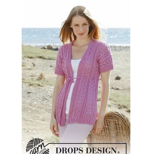 May include: A pink knitted cardigan with a V-neck and short sleeves. The cardigan has a tie at the waist and a lacy pattern. The model is wearing the cardigan with white pants and is standing in front of a beach.
