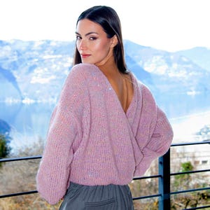 May include: A soft, chunky knit jumper in a light lavender pink hue with subtle multicoloured flecks throughout. The garment features a deep V-neckline that crosses over at the back, long sleeves with ribbed cuffs, and a relaxed, loose-fitting silhouette.