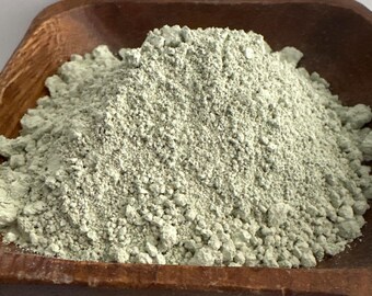 May include: A close-up shot of a pile of fine, pale green powder in a wooden bowl. The powder has a slightly granular texture and appears to be a cosmetic or skincare ingredient. The bowl is a warm brown color, providing a contrast to the powder.