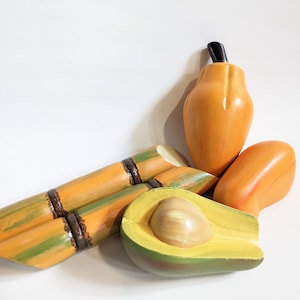 Könnte beinhalten: Eine Sammlung handbemalter Holzfrüchte, darunter eine Papaya mit schwarzem Stiel, eine halbierte Avocado und ein Zuckerrohrstück. Die Früchte sind in Orange-, Grün- und Brauntönen bemalt.