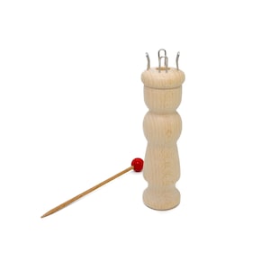 Könnte beinhalten: Ein Strickliesel aus Holz mit Metallhaken und eine Holzstricknadel mit einer roten Perle. Die Strickliesel ist hell und hat eine abgerundete Form. Die Nadel ist braun und spitz. Die rote Perle ist klein und rund. Zum Stricken.