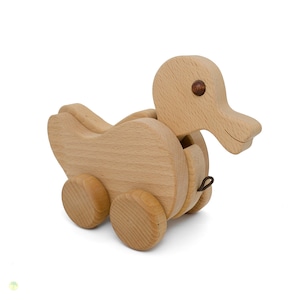 Può includere: Un'anatra di legno giocattolo con le ruote. L'anatra è fatta di legno marrone chiaro e ha un occhio marrone. L'anatra è su uno sfondo bianco.