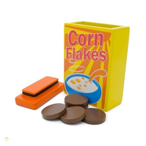Puede incluir: Una caja amarilla con las palabras "Corn Flakes" impresas en ella. La caja está abierta y contiene varios discos de chocolate marrón. También hay un pequeño objeto rectangular naranja y negro al lado de la caja.