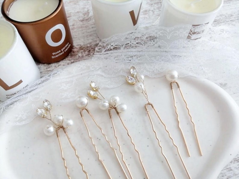 May include: Six gold hair pins with pearl and crystal accents. The pins are arranged on a white surface.