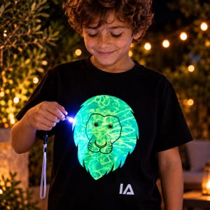 Könnte beinhalten: Ein schwarzes Kurzarm-T-Shirt mit einer großen, leuchtend grünen Löwenkopf-Grafik auf der Brust. Das Design wird durch ein kleines, in der Hand gehaltenes Lichtwerkzeug beleuchtet, und die Buchstaben IA sind weiß am unteren Saum aufgedruckt.