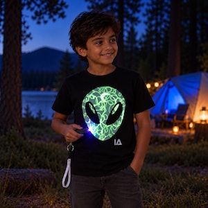 May include: A black short-sleeved t-shirt featuring a large, glowing green alien head graphic on the chest. The alien design is filled with a chaotic scribble pattern and a small crescent moon shape. The letters IA are printed in white below the graphic.