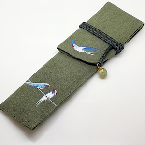Könnte beinhalten: Ein grünes Stoff-Stift-Etui mit Klappenverschluss und einer schwarzen Schnur mit einer jadegrünen Perle. Das Etui ist mit gestickten blauen Vögeln auf einem weißen Draht verziert.