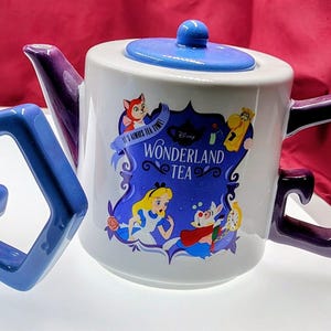 May include: A white ceramic mug and teapot set featuring Alice in Wonderland characters. The mug has a blue interior and handle, while the teapot has a blue lid and purple handle. The teapot reads "Wonderland Tea".