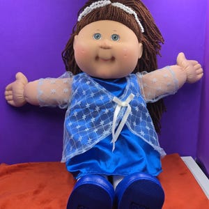 Könnte beinhalten: Eine Puppe mit braunen Wollhaaren, einem weißen Stirnband, blauen Augen und rosigen Wangen. Die Puppe trägt ein blaues Satinkleid mit einem transparenten weißen Überwurf mit Schneeflockenmuster und einer weißen Schleife sowie blauen Schuhen.