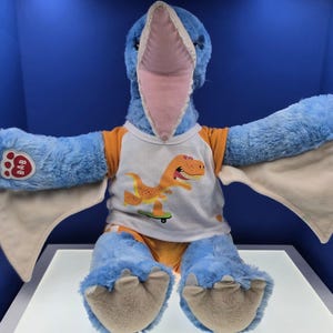May include: A blue plush pterodactyl toy with a pink beak and tan wing undersides. It wears a white t-shirt with orange sleeves featuring an orange dinosaur on a skateboard. The right paw has a red B-A-B logo patch. The feet have tan claws.