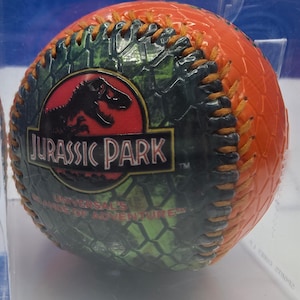 May include: An orange baseball with a textured surface and brown stitching. The ball features a green and black dinosaur skin pattern with the Jurassic Park logo and the text Universal's Islands of Adventure printed on the front surface.
