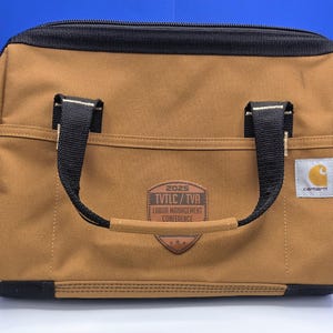 May include: A tan Carhartt lunch bag with black trim and handles. The front features a brown leather-like shield patch with the text 2025 TVTLC / TVA LABOR MANAGEMENT CONFERENCE and a small white Carhartt logo tag on the right side of the pocket.