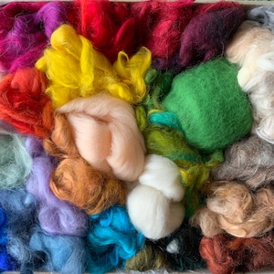 Könnte beinhalten: Eine Kiste voller farbenfroher Wollvliese in verschiedenen Farbtönen. Die Auswahl umfasst leuchtende Farben wie Rot, Rosa, Gelb, Grün, Blau und Braun. Die Wolle ist weich und flauschig, ideal für Textilkunstprojekte.