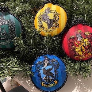 May include: Four round Christmas ornaments with crests representing Hogwarts houses: Slytherin, Hufflepuff, Gryffindor, and Ravenclaw. Each ornament has a black rope trim and a silver loop for hanging. The ornaments are displayed on a bed of green pine needles.