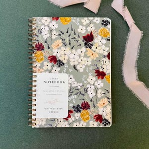 Op de afbeelding: Een spiraalgebonden notitieboek met een bloemenpatroon in wit, geel en bordeauxrood op een saliegroene achtergrond. Het notitieboek heeft een label met de tekst "Lined Notebook, 100 pages, handcrafted in Whitney's, Tennessee studio, Whitney Rain Studio."