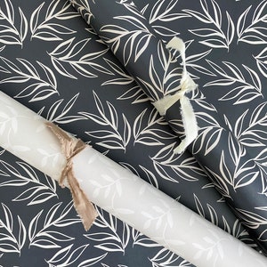 Puede incluir: Dos rollos de papel de regalo con un patr&oacute;n de hojas blancas sobre un fondo azul oscuro. Los rollos est&aacute;n atados con una cinta blanca y marr&oacute;n.