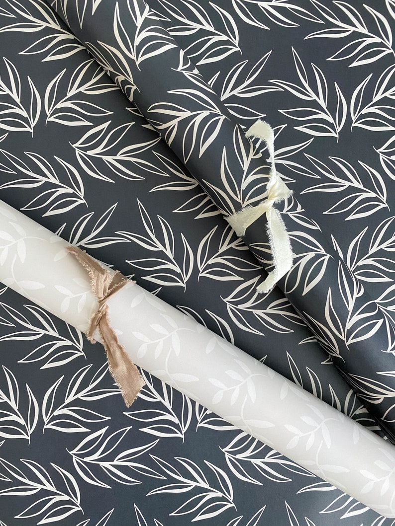 Puede incluir: Dos rollos de papel de regalo con un patr&oacute;n de hojas blancas sobre un fondo azul oscuro. Los rollos est&aacute;n atados con una cinta blanca y marr&oacute;n.