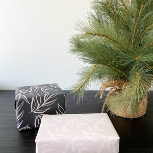 Puede incluir: Dos cajas de regalo envueltas en papel blanco y negro con un patr&oacute;n de hojas. Las cajas est&aacute;n sobre una superficie negra delante de un peque&ntilde;o pino verde.