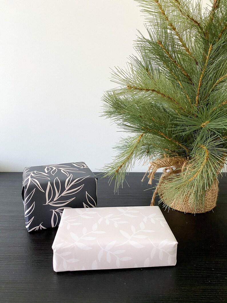 Puede incluir: Dos cajas de regalo envueltas en papel blanco y negro con un patr&oacute;n de hojas. Las cajas est&aacute;n sobre una superficie negra delante de un peque&ntilde;o pino verde.
