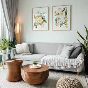 May include: A living room scene featuring a gray sofa with decorative pillows and a patterned throw. Two framed floral watercolor paintings hang above the sofa. A wooden coffee table and side table, along with a woven pouf, complete the decor.