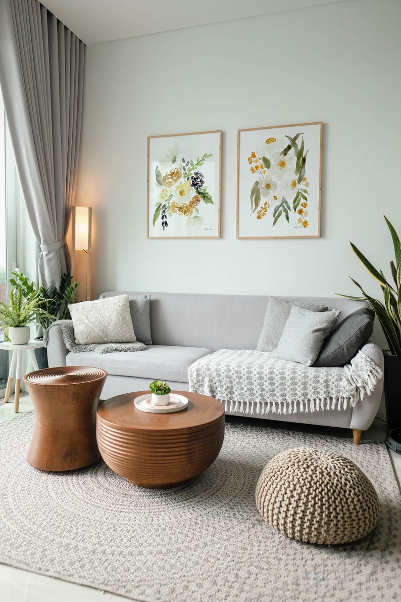 May include: A living room scene featuring a gray sofa with decorative pillows and a patterned throw. Two framed floral watercolor paintings hang above the sofa. A wooden coffee table and side table, along with a woven pouf, complete the decor.