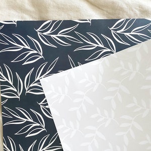 Puede incluir: Dos hojas de papel de regalo con un patr&oacute;n de hojas blancas. Una hoja es negra y la otra es blanca.