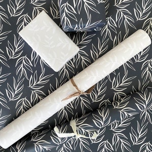 Puede incluir: Un rollo de papel de regalo con estampado blanco y negro con una caja de regalo blanca y una caja de regalo blanca m&aacute;s peque&ntilde;a con estampado blanco y negro. El estampado es de hojas blancas sobre un fondo negro.