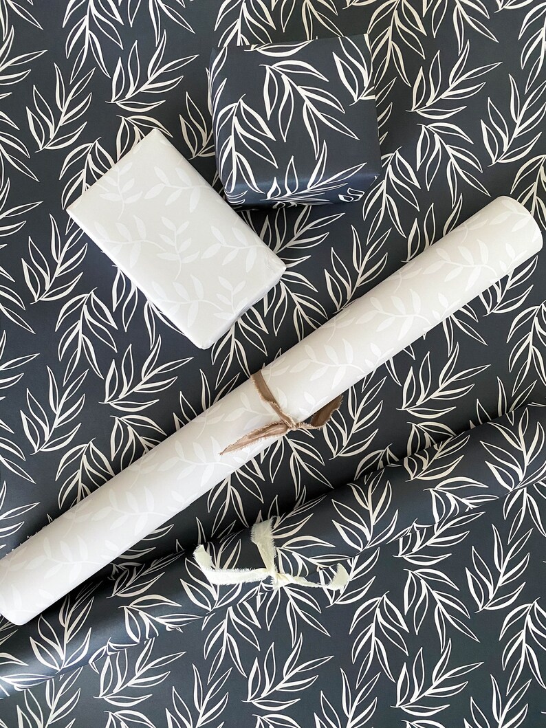 Puede incluir: Un rollo de papel de regalo con estampado blanco y negro con una caja de regalo blanca y una caja de regalo blanca m&aacute;s peque&ntilde;a con estampado blanco y negro. El estampado es de hojas blancas sobre un fondo negro.