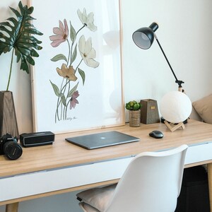 May include: A light wood desk with a white drawer, a laptop, a black speaker, a camera, a framed watercolor print of flowers, a lamp with a white moon-shaped lampshade, and a white chair with a light brown cushion.